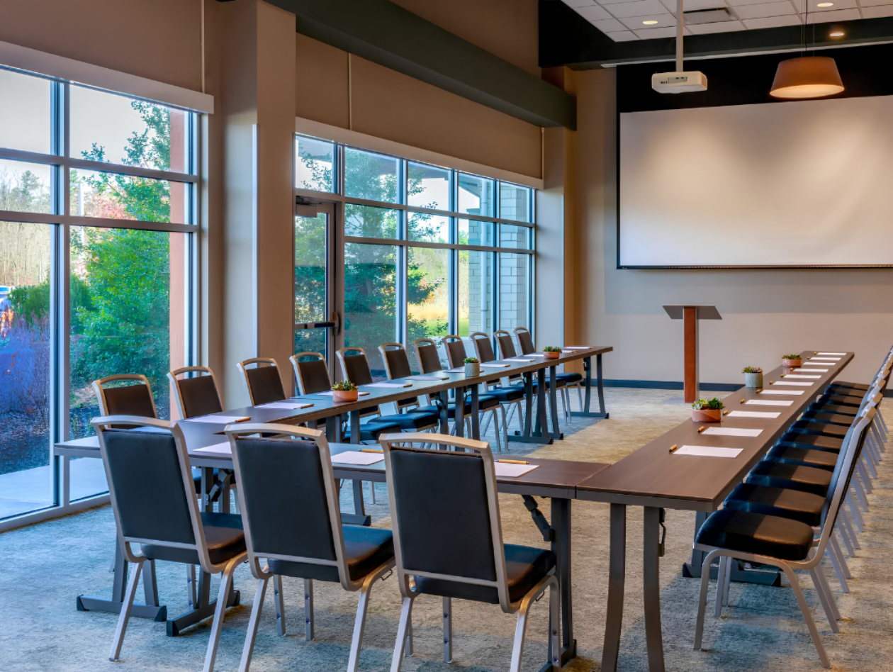 Modern conference room with two long tables arranged in a U-shape, chairs, a lectern, notepads, and large windows showing greenery outside.