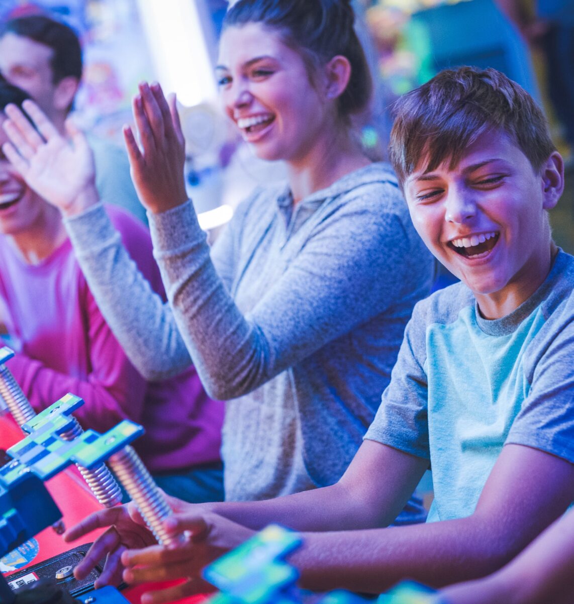 Four kids laugh and play an arcade game together, each holding game controllers, surrounded by colorful lights and machines.