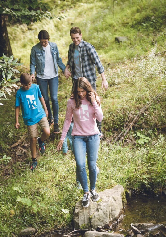 Four people walk down a grassy hill near a creek; a woman leads, followed by two children and a man. The group looks focused on their steps.