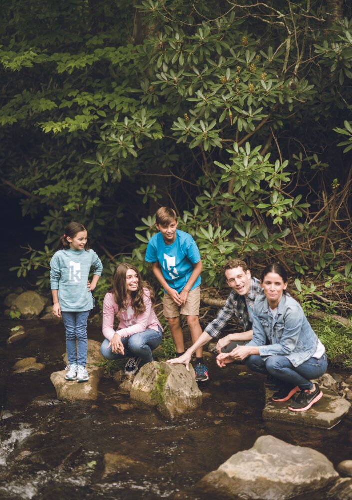 A family of five poses on rocks in a creek surrounded by green trees, smiling and enjoying nature together.