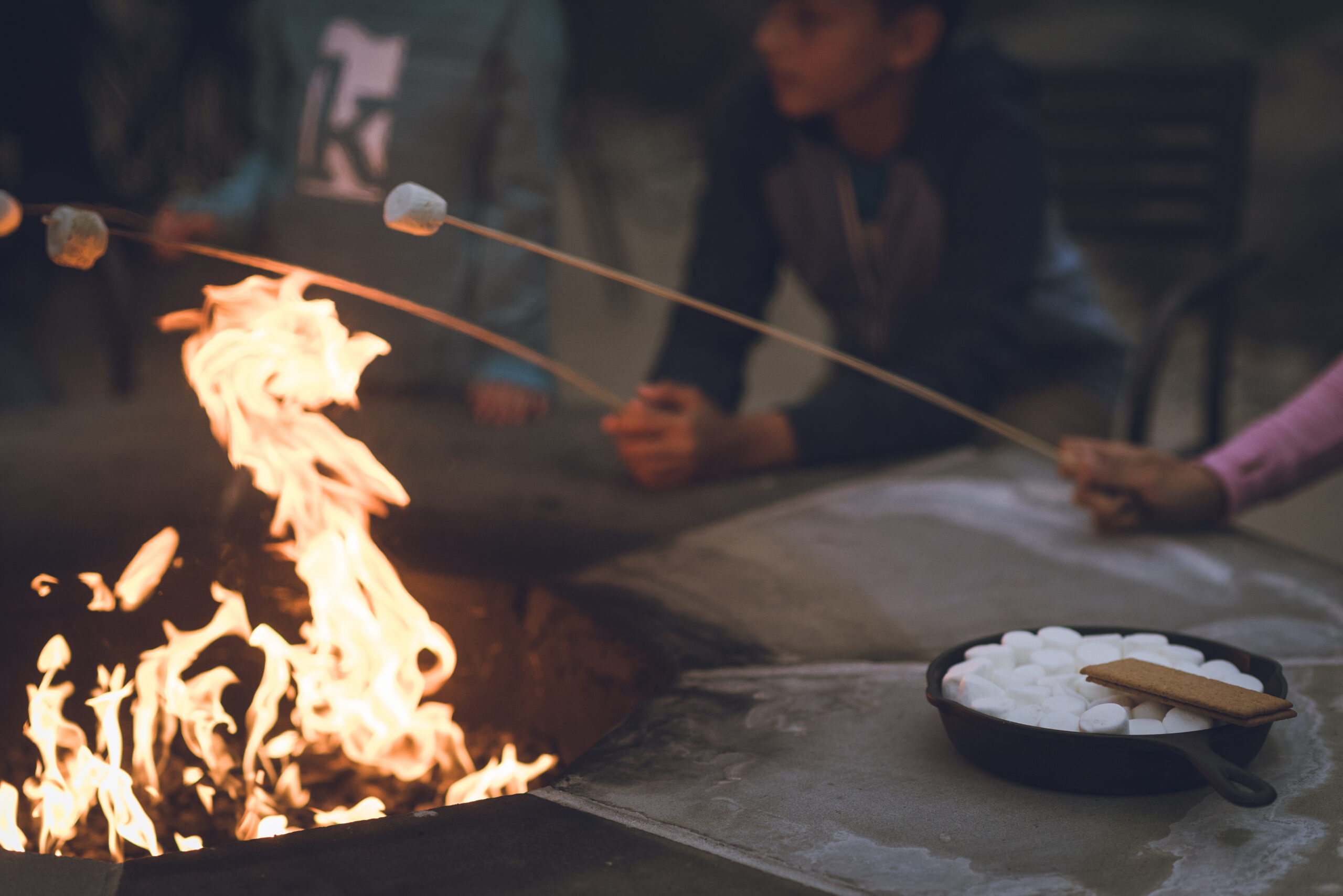 People roast marshmallows over a fire pit, with a skillet of marshmallows and graham crackers on the table nearby.