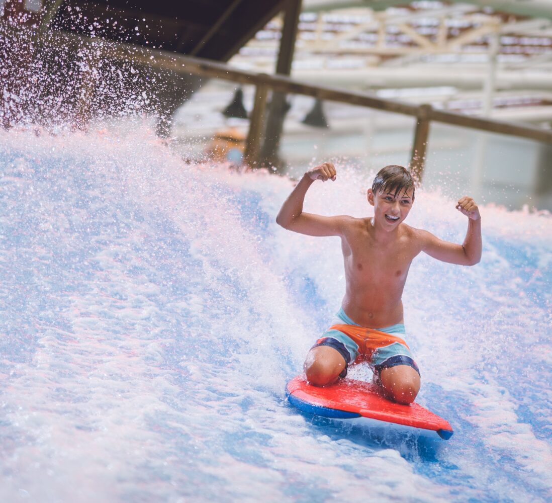 A boy in swim trunks rides a wave on a red bodyboard indoors, smiling and flexing his arms while water splashes around him.