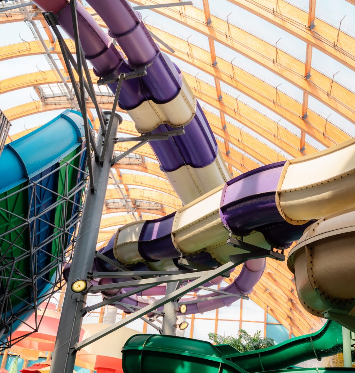 Curving indoor water slides in blue, purple, tan, and green, with a wooden arched ceiling above and sunlight streaming through large windows.