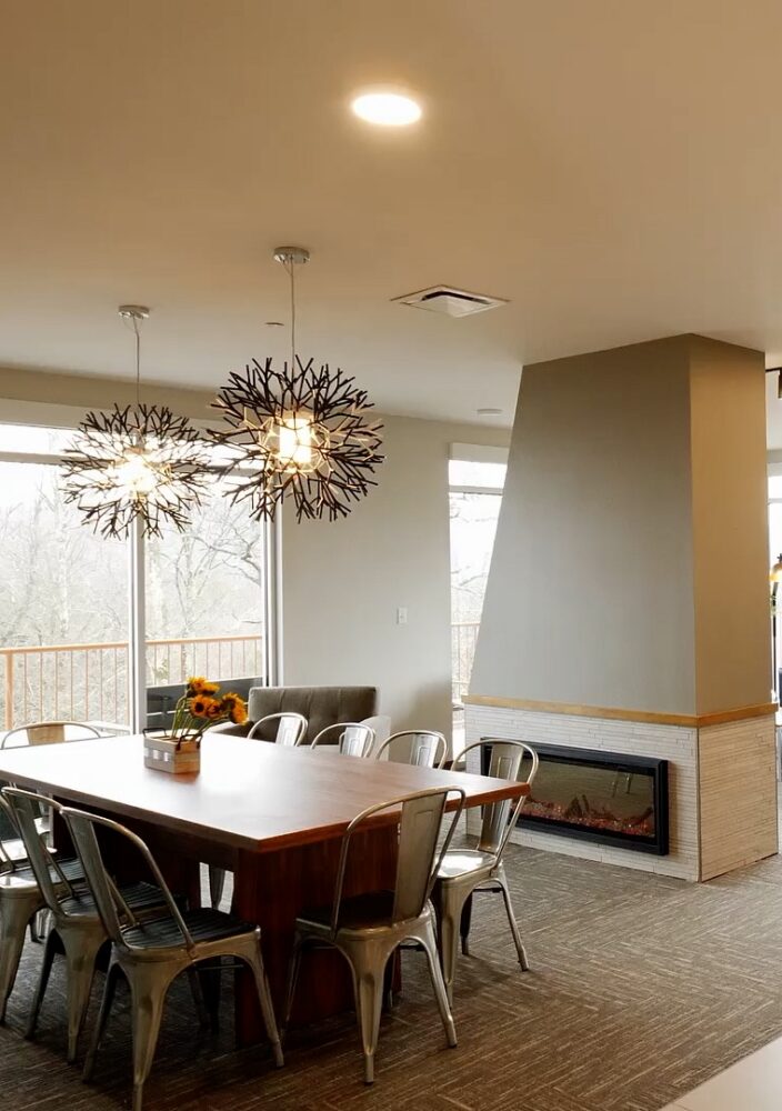 Modern open kitchen and dining area with unique pendant lights, a wooden table, metal chairs, and a cozy living space with large windows in the background.