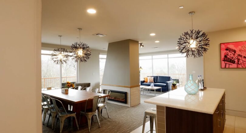 Modern open kitchen and dining area with unique pendant lights, a wooden table, metal chairs, and a cozy living space with large windows in the background.