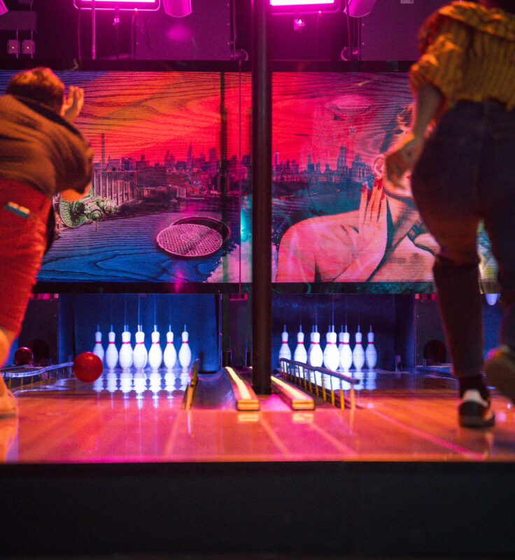Two people bowl side by side in a neon-lit alley, colorful mural and glowing pins visible ahead.
