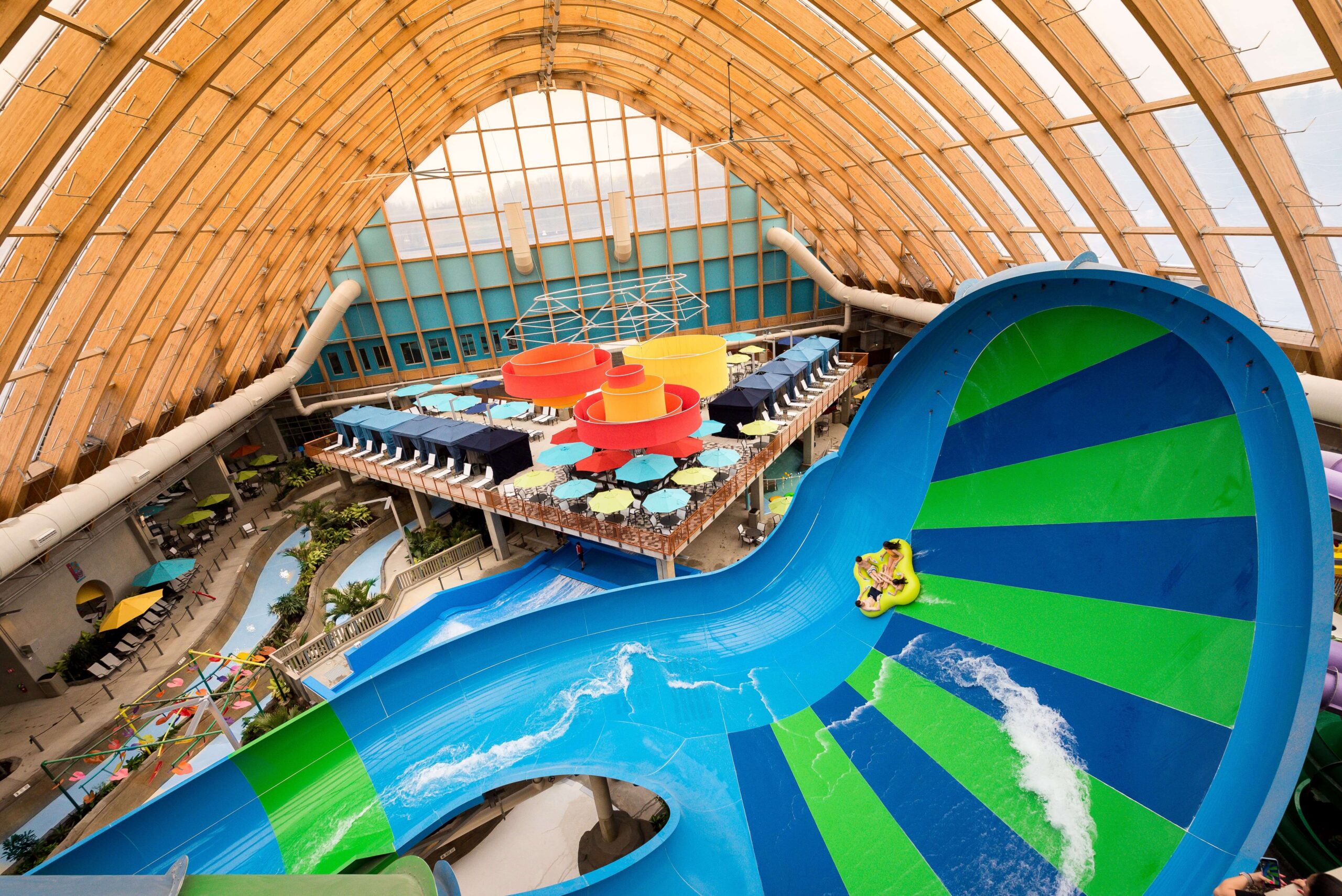 Indoor water park with a large, curving green and blue water slide, riders on a raft, and tables with umbrellas below a wooden arched roof.