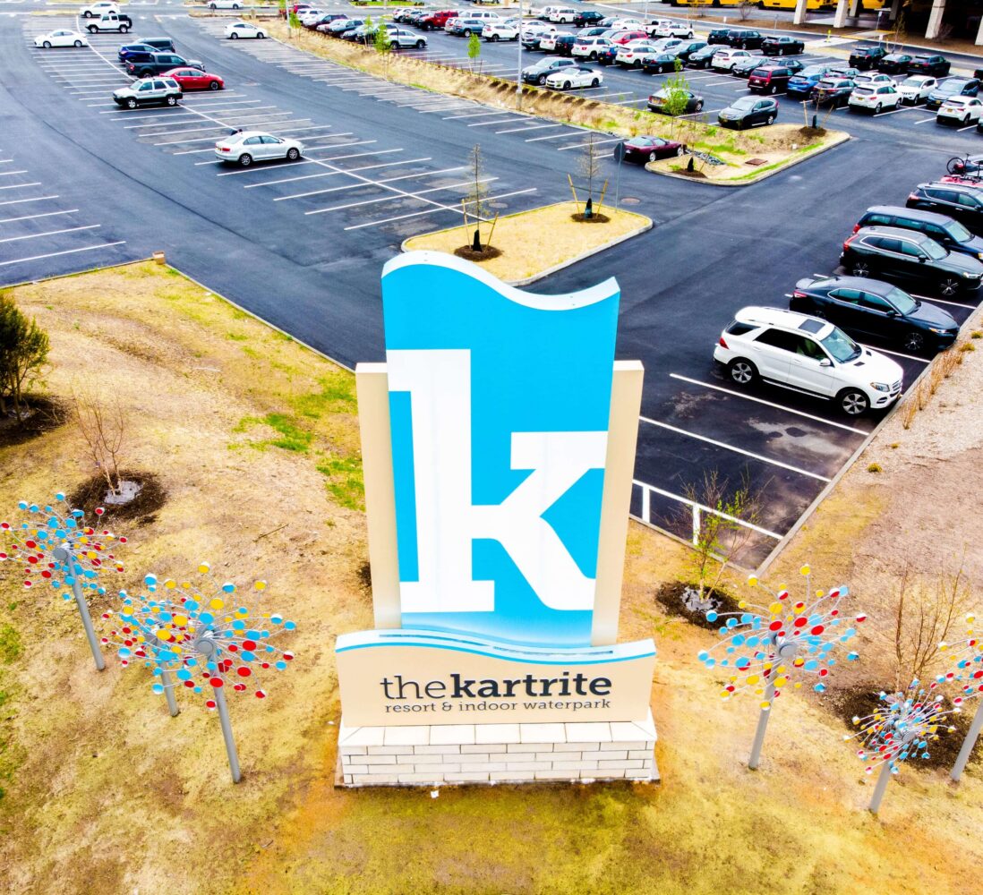 A large blue and white k sign for The Kartrite Resort & Indoor Waterpark stands in a parking lot with cars and sparse trees.