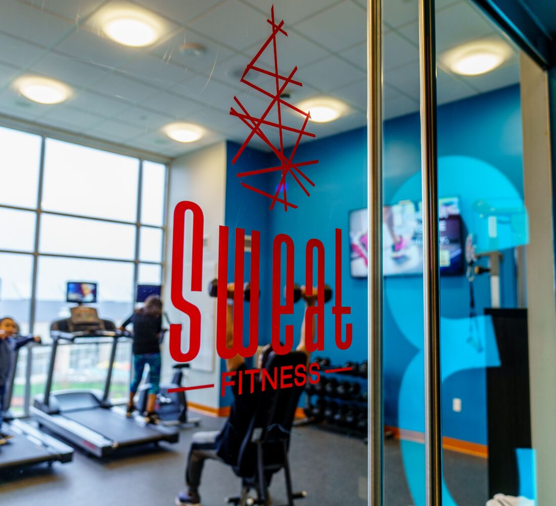 Red Sweat Fitness logo on a glass door, with people using treadmills and weight equipment in a bright gym interior behind it.