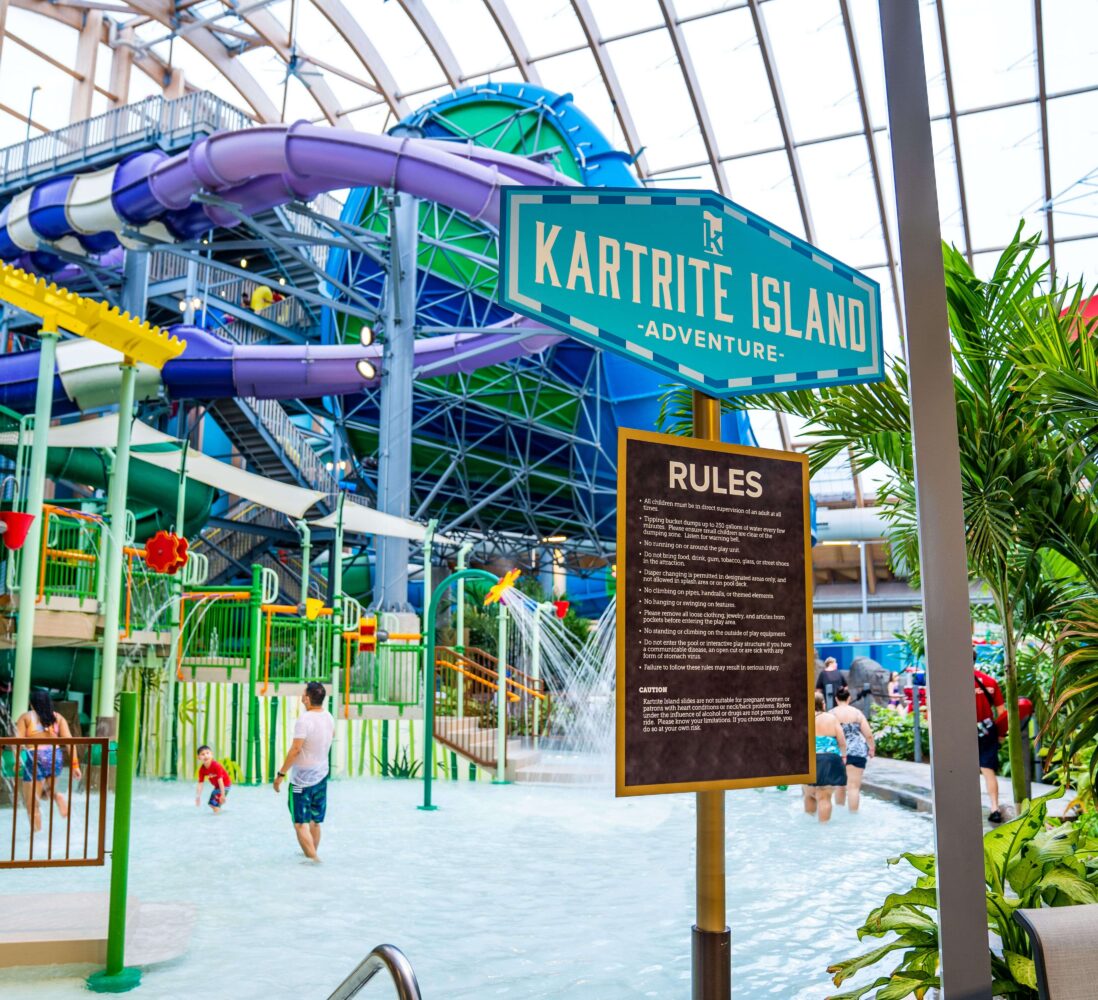 Indoor water park with people playing, colorful slides, a Kartrite Island sign, and a rules board under a large glass roof with tropical plants.