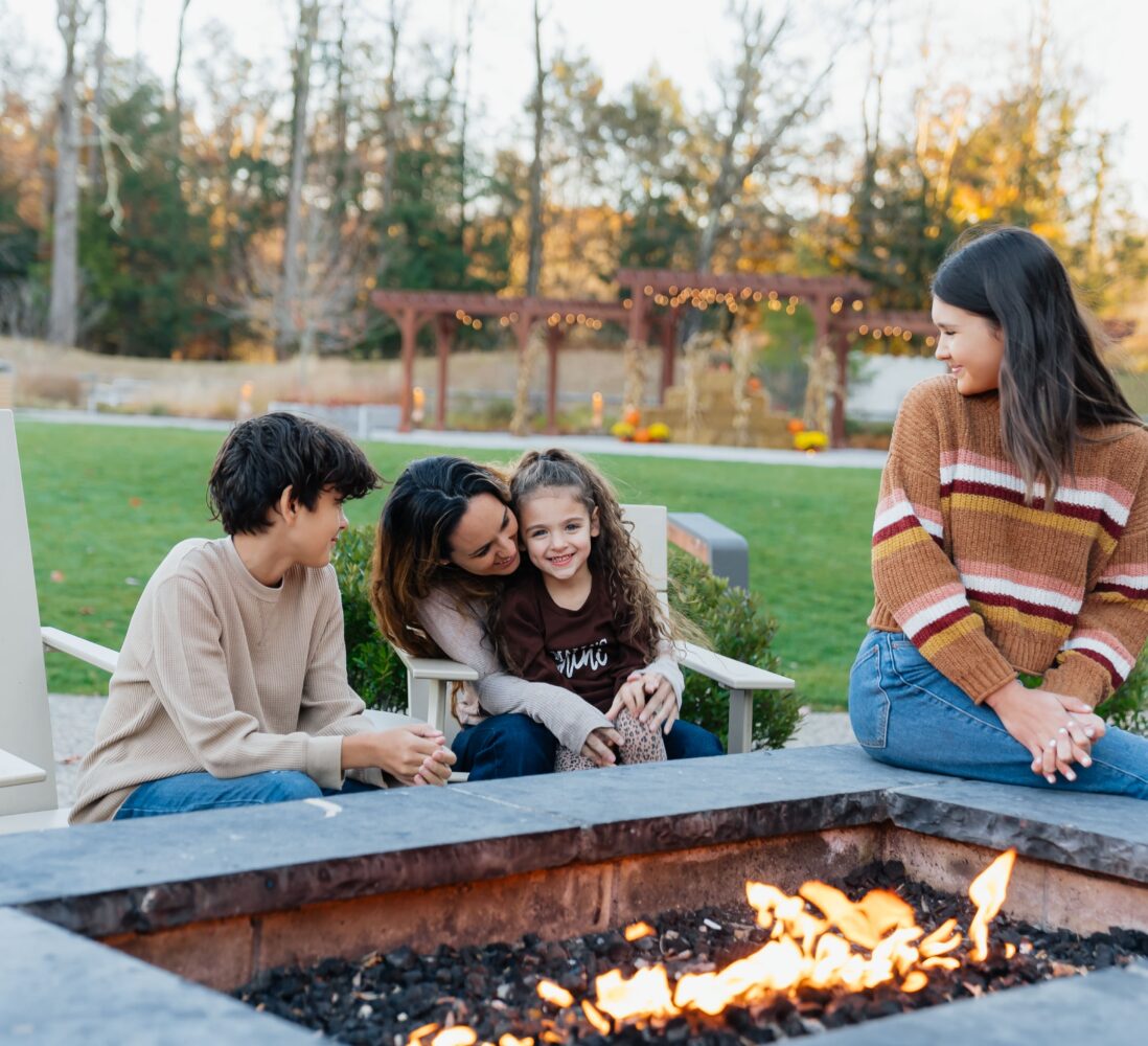 A family of four sits by an outdoor fire pit, smiling and enjoying time together in a backyard with trees and string lights in the background.