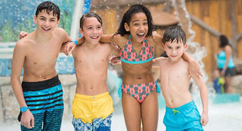Four smiling kids in swimwear stand arm-in-arm at a water park, with splashing water behind them.