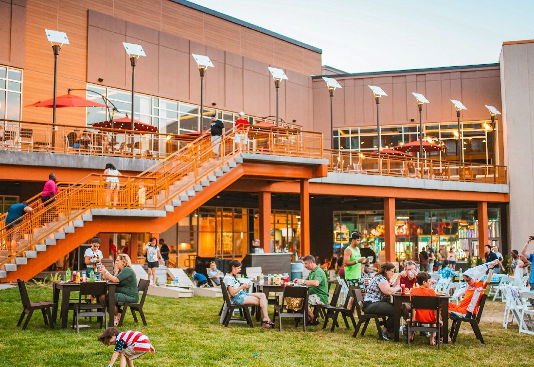 People dine and gather outdoors on a grassy lawn by a modern building with large windows and an upper deck, enjoying a lively, sunny atmosphere.
