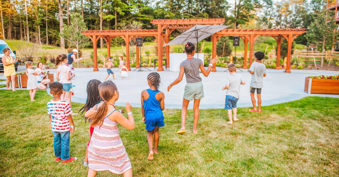 Children play and dance together on grass near a wooden pergola in a sunny outdoor park setting surrounded by trees.