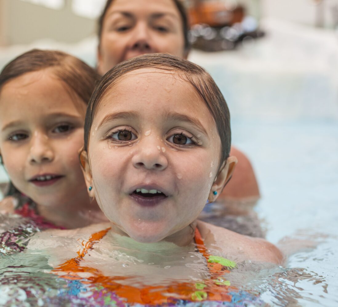 Two young girls in swimsuits smile while swimming in a pool, with an adult behind them. Their faces are wet and they appear to be having fun.