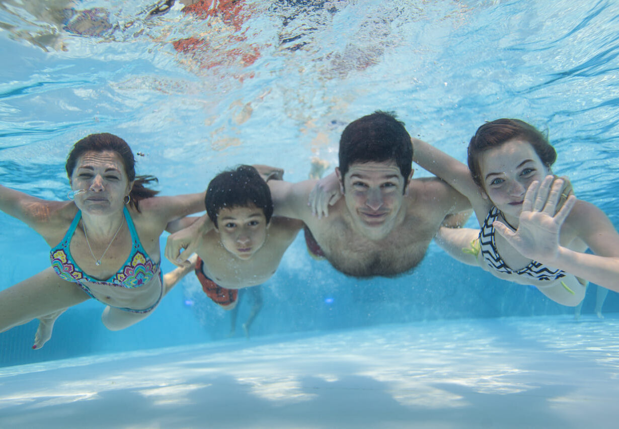 Four people smile underwater in a swimming pool, with one person waving at the camera. Sunlight reflects on the water.