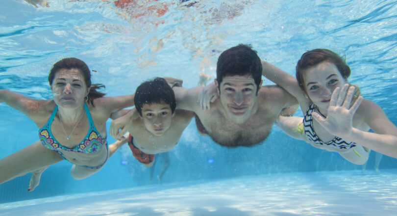 Four people smile underwater in a swimming pool, with one person waving at the camera. Sunlight reflects on the water.