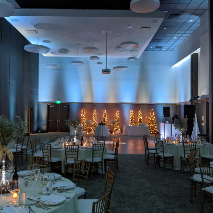Elegant banquet hall with round tables, white linens, candles, and string-lit trees at the back, creating a warm, festive atmosphere.