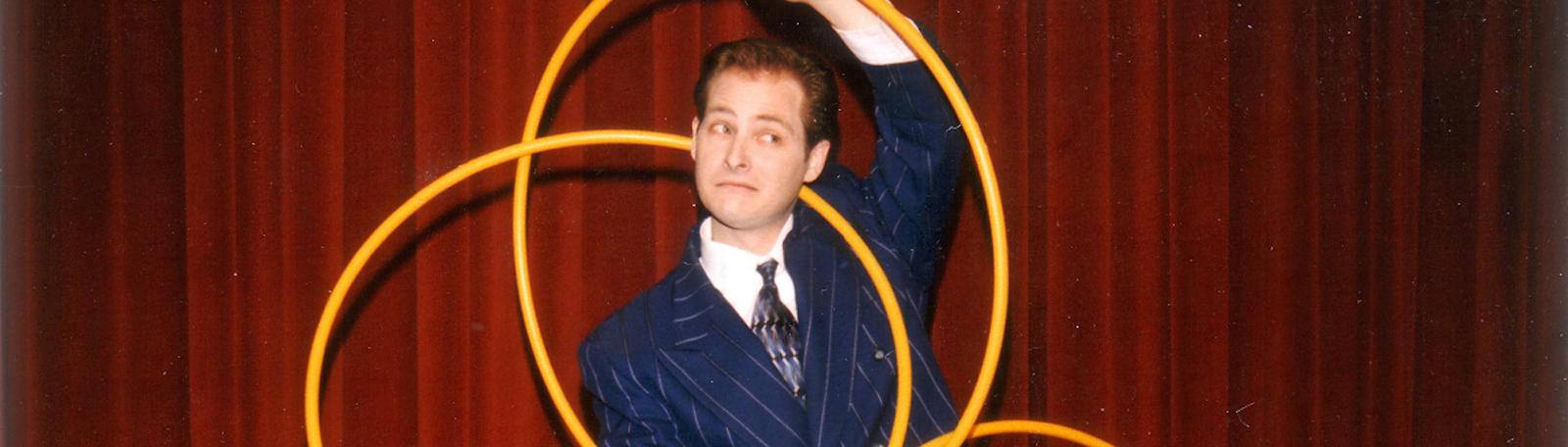 Man in a suit performs with several yellow hoops in front of a red curtain, looking off to the side with a playful expression.