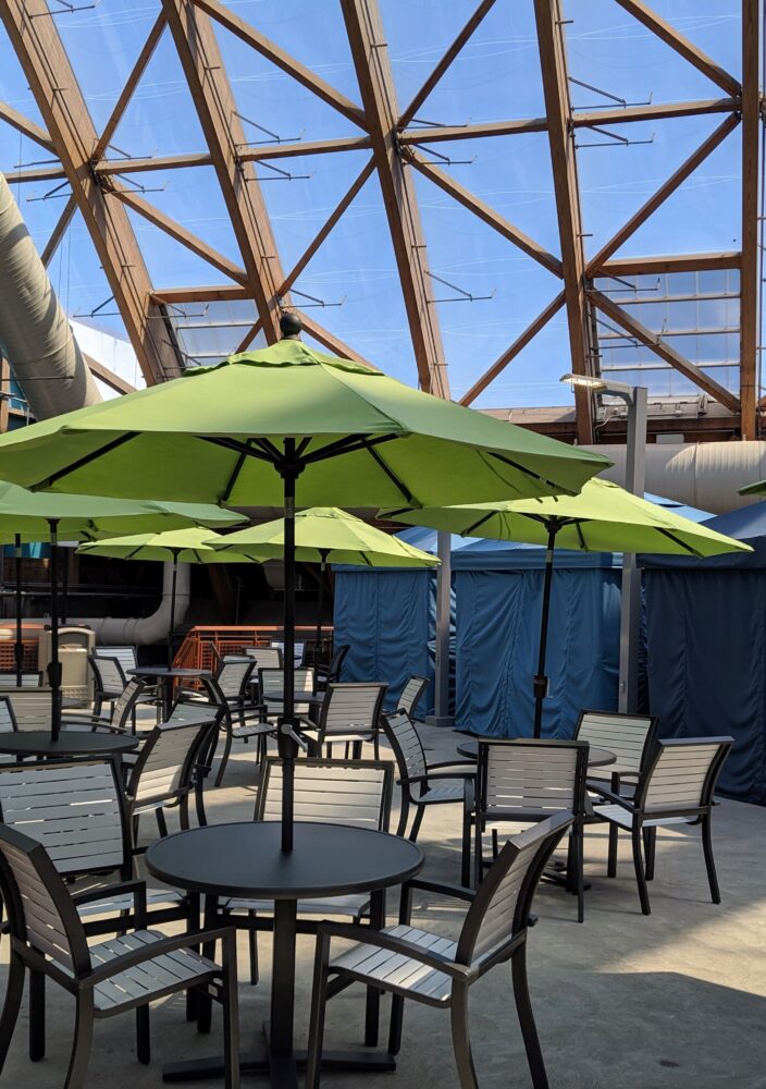Outdoor seating area with black tables, chairs, and green umbrellas under a glass roof structure with blue walls and large pipes.