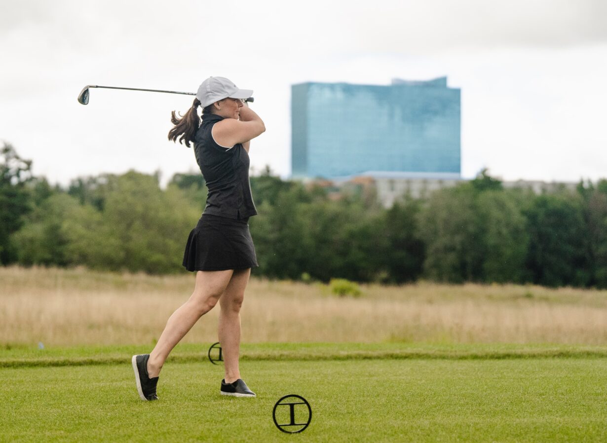 A woman in a black outfit and white cap swings a golf club on a grassy course with trees and a tall blue building in the background.