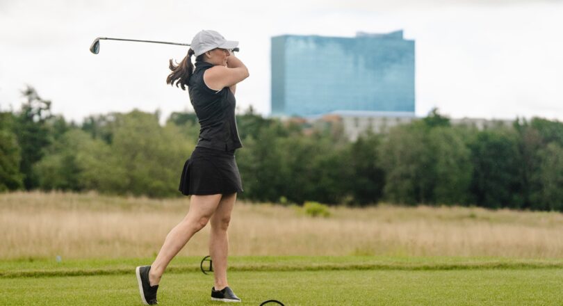 A woman in a black outfit and white cap swings a golf club on a grassy course with trees and a tall blue building in the background.