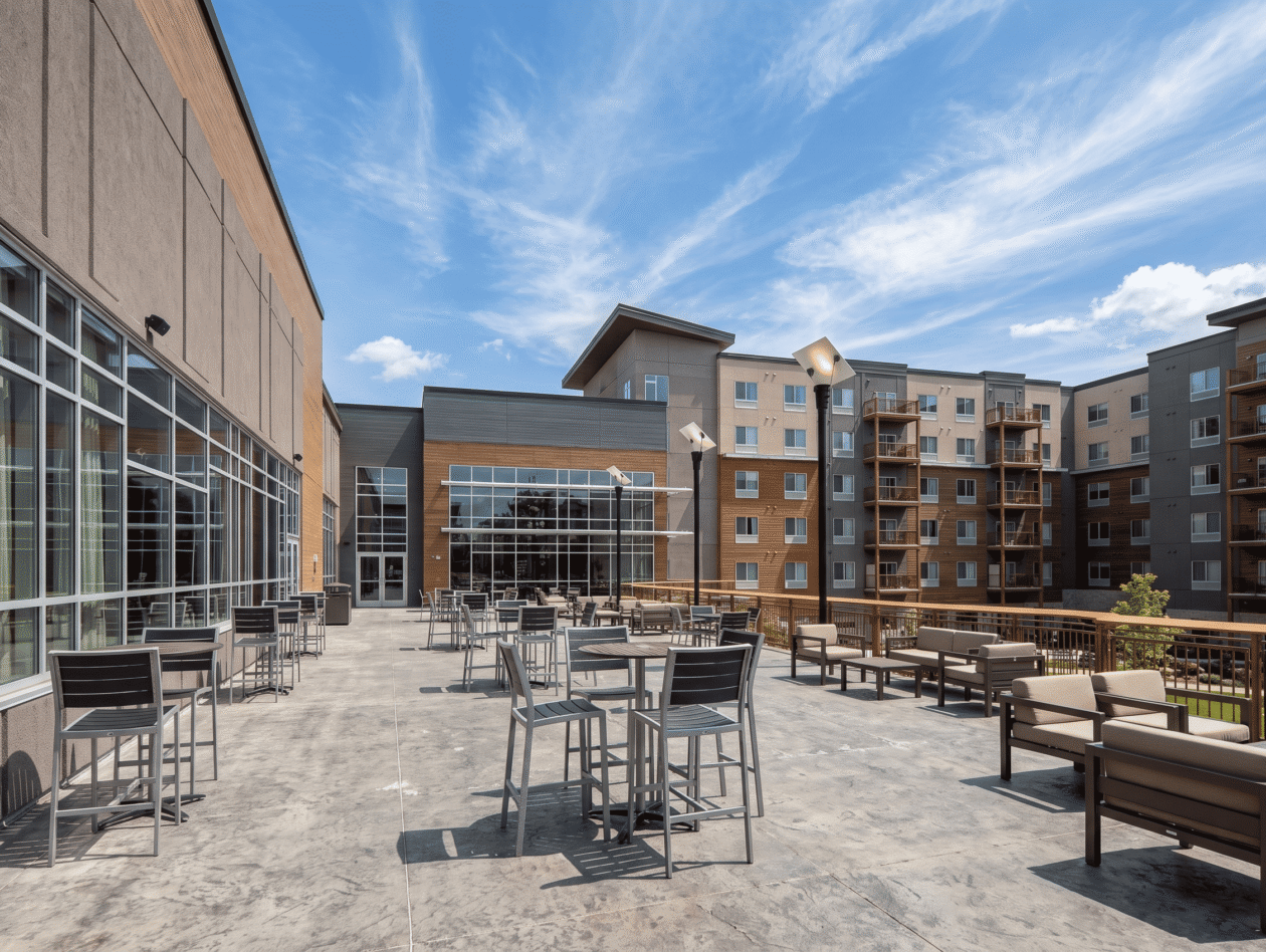 Outdoor patio with tables and chairs, overlooking a modern apartment building under a blue sky with wispy clouds.