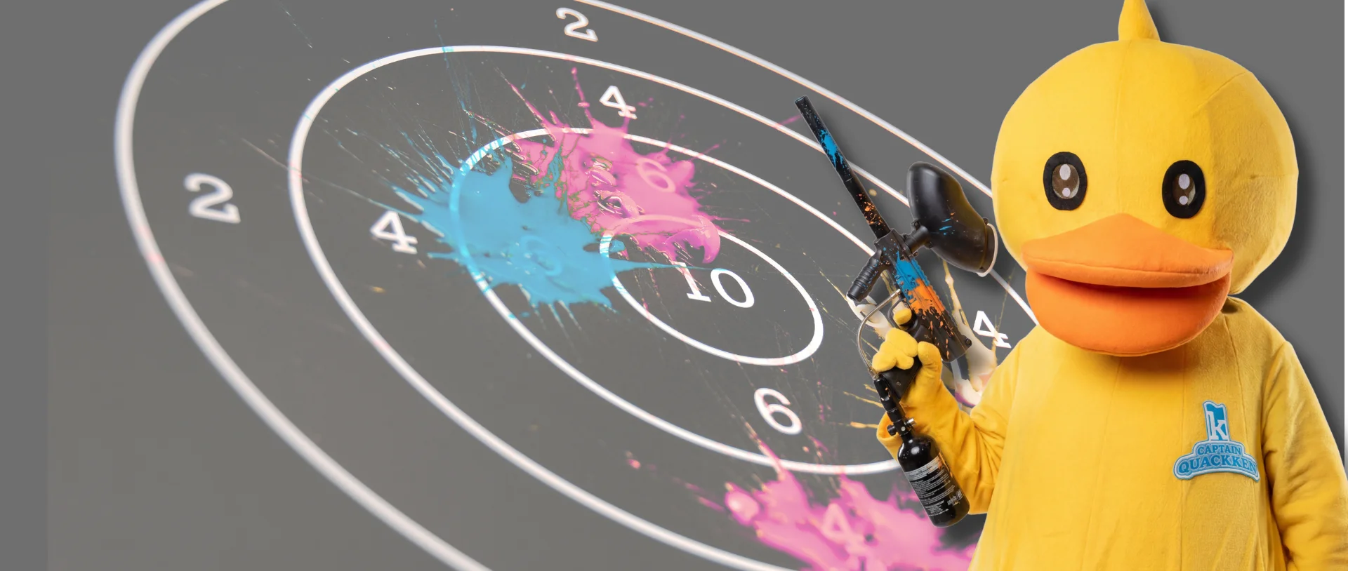 A person in a yellow duck costume holds a paintball gun in front of a target splattered with pink and blue paint.