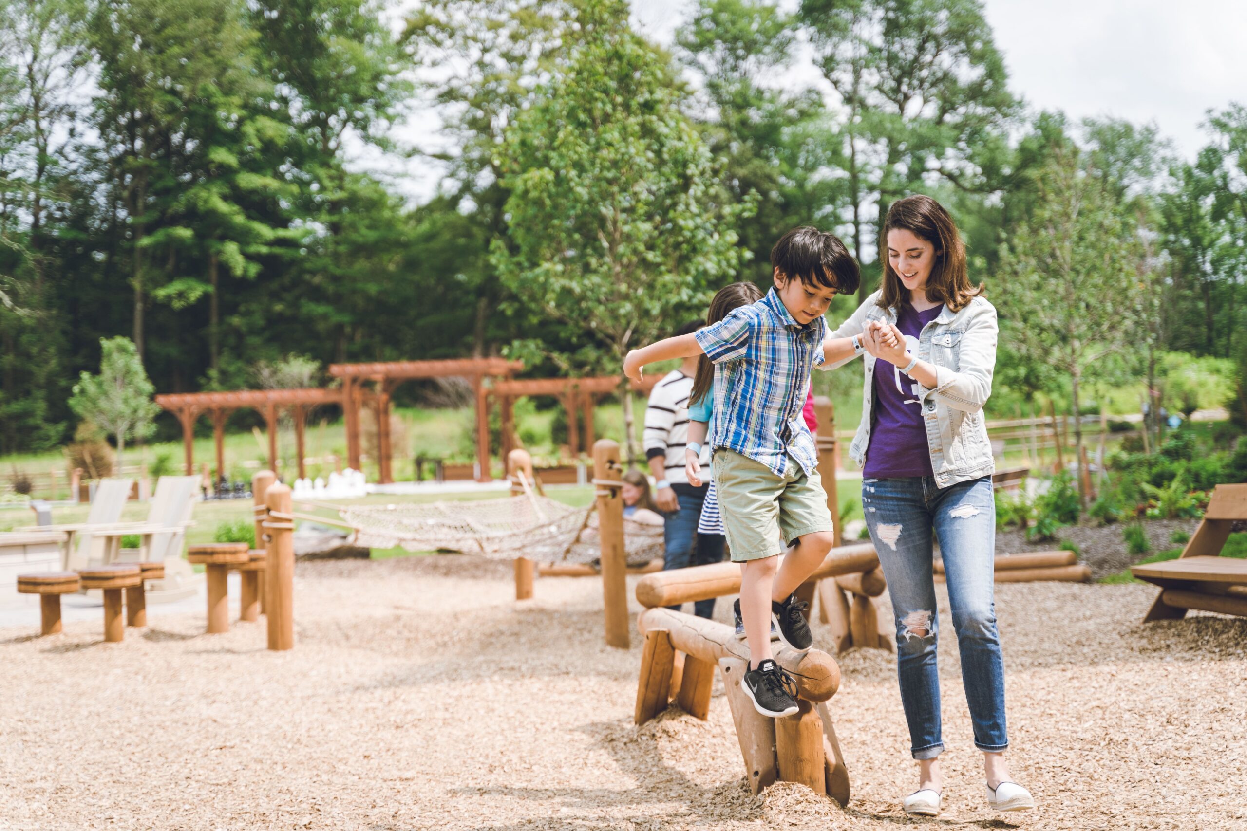 A child balances on a wooden beam in a playground while an adult holds their hand, surrounded by trees and outdoor seating.