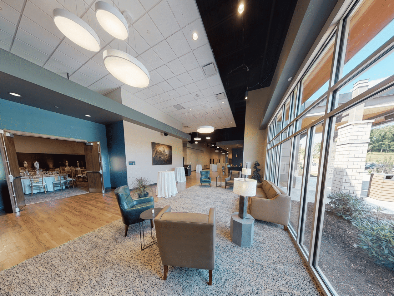 Modern lobby with seating, large windows, and round ceiling lights. Open doors reveal a banquet room with tables and chairs in the background.