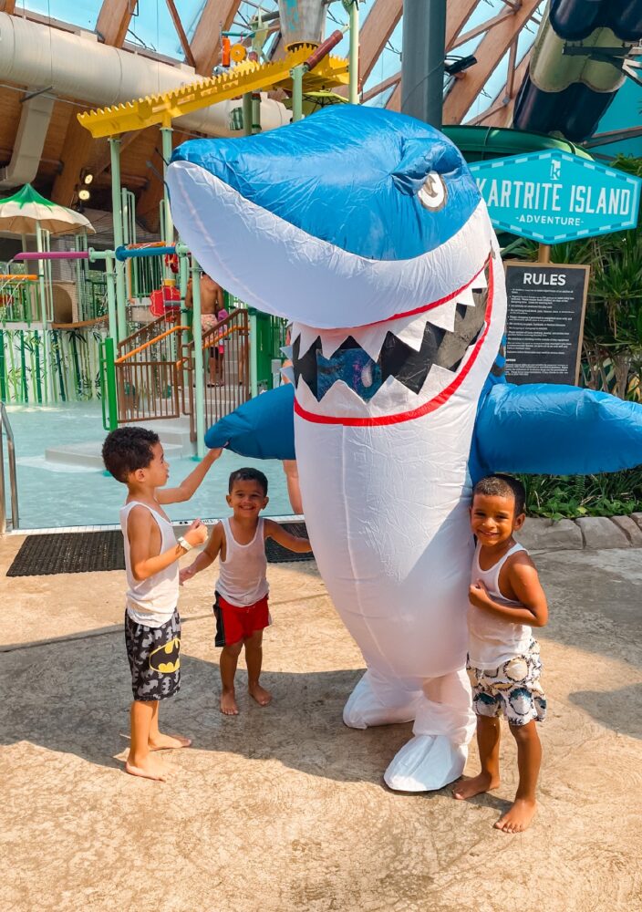 Three kids in swimwear smile and pose with a person in a large inflatable shark costume at an indoor water park.