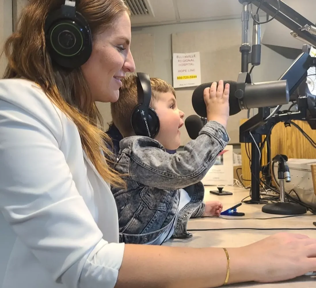 A woman and a young boy with headphones sit at a radio studio desk; the boy excitedly speaks into a microphone.