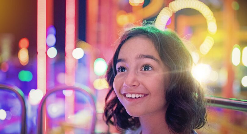 Smiling child with short dark hair enjoys colorful, bright lights at an amusement park, glowing with excitement.