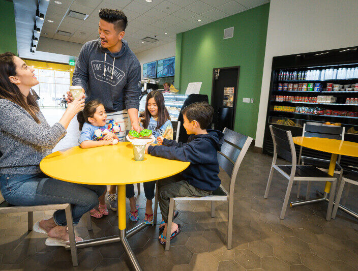 A family of five enjoys ice cream together at a yellow table in a modern shop with green walls and a fridge stocked with drinks.