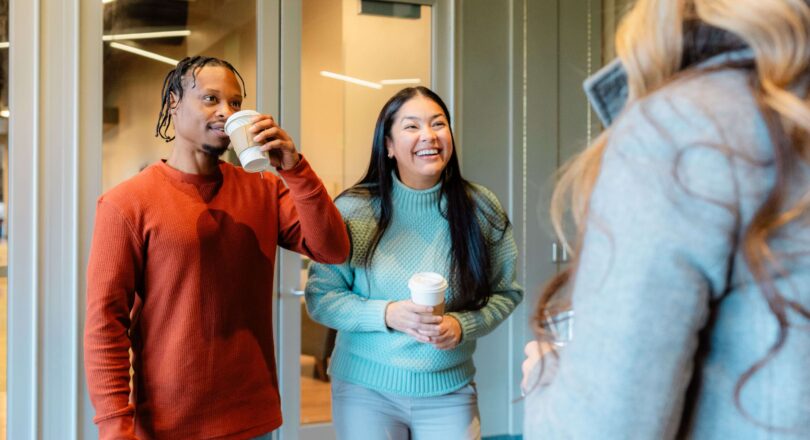 Two people smiling and talking while holding coffee cups, facing a third person whose back is to the camera, in an indoor setting.