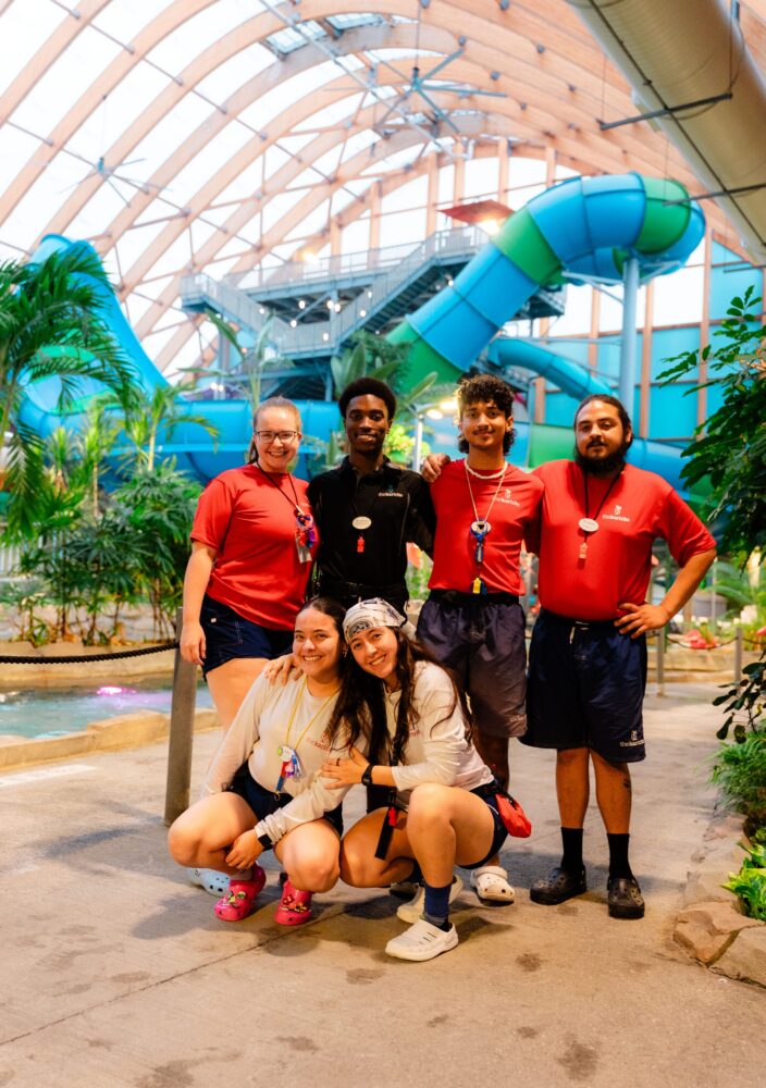 Six lifeguards pose and smile inside an indoor water park, with waterslides, tubes, and tropical plants visible in the bright, spacious background.