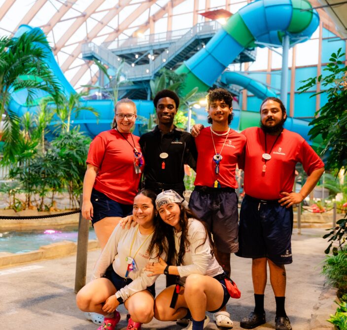 Six lifeguards pose and smile inside an indoor water park, with waterslides, tubes, and tropical plants visible in the bright, spacious background.