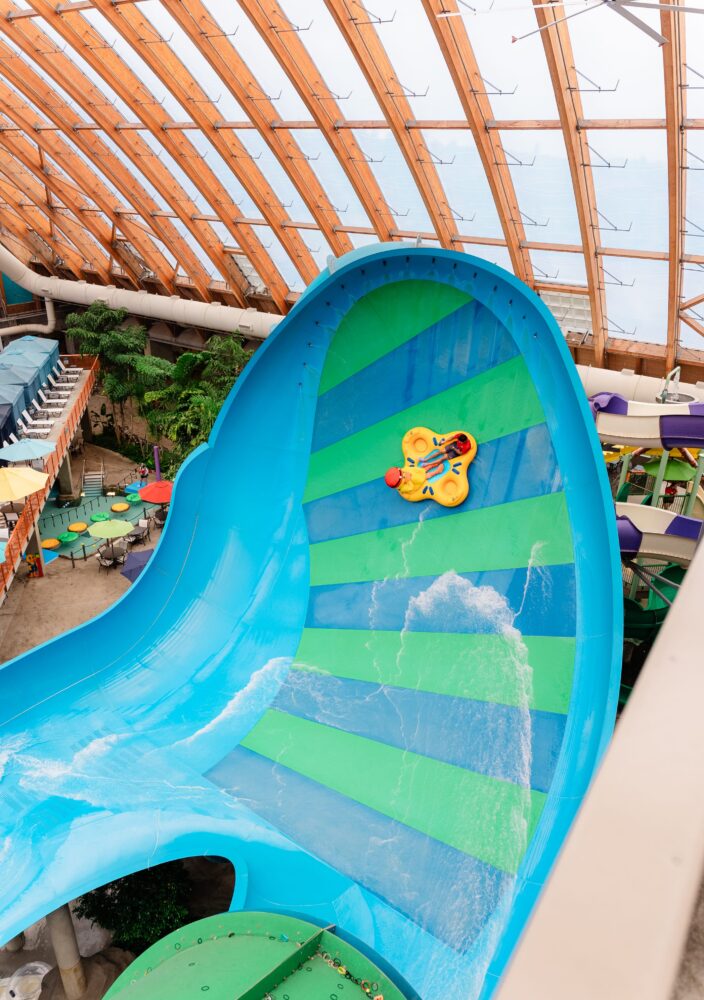 Two people ride a yellow float down a steep, striped green and blue water slide inside an indoor water park with a wooden arched roof.