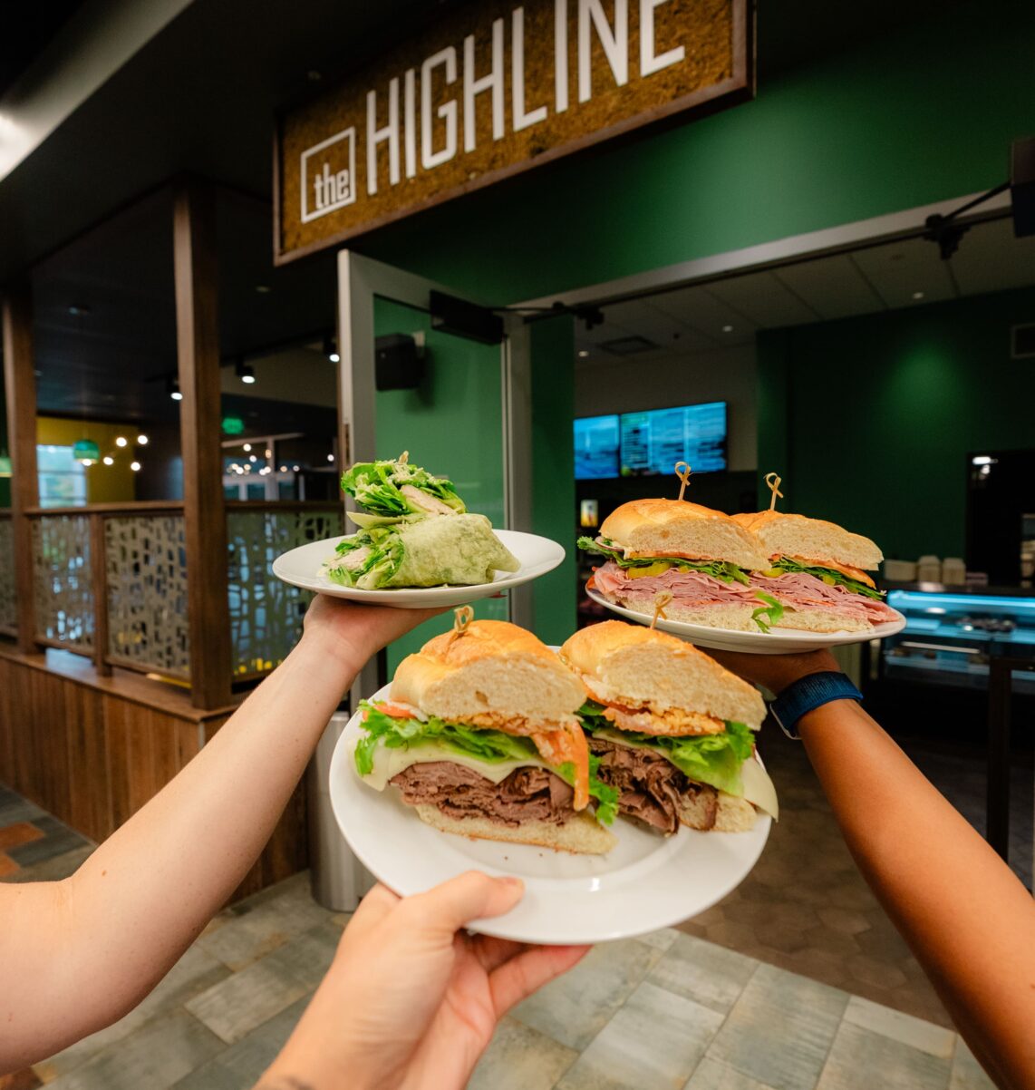Three hands hold plates of sandwiches and salad in front of a sign that reads the HIGHLINE in a modern, green-themed restaurant.