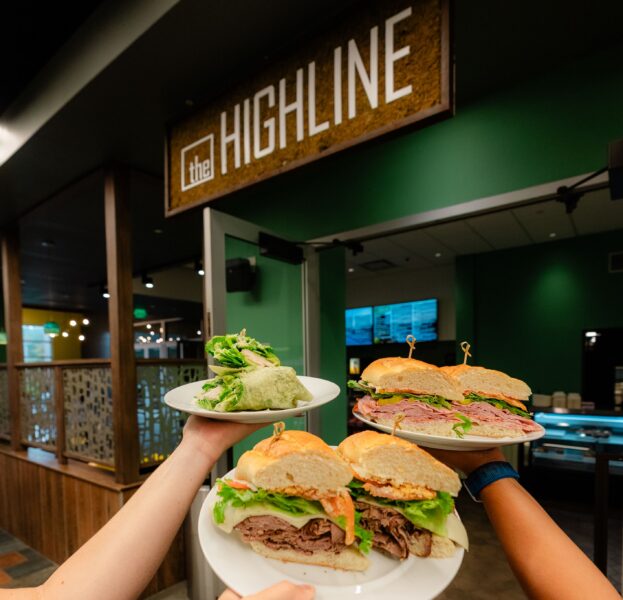 Three hands hold plates of sandwiches and salad in front of a sign that reads the HIGHLINE in a modern, green-themed restaurant.