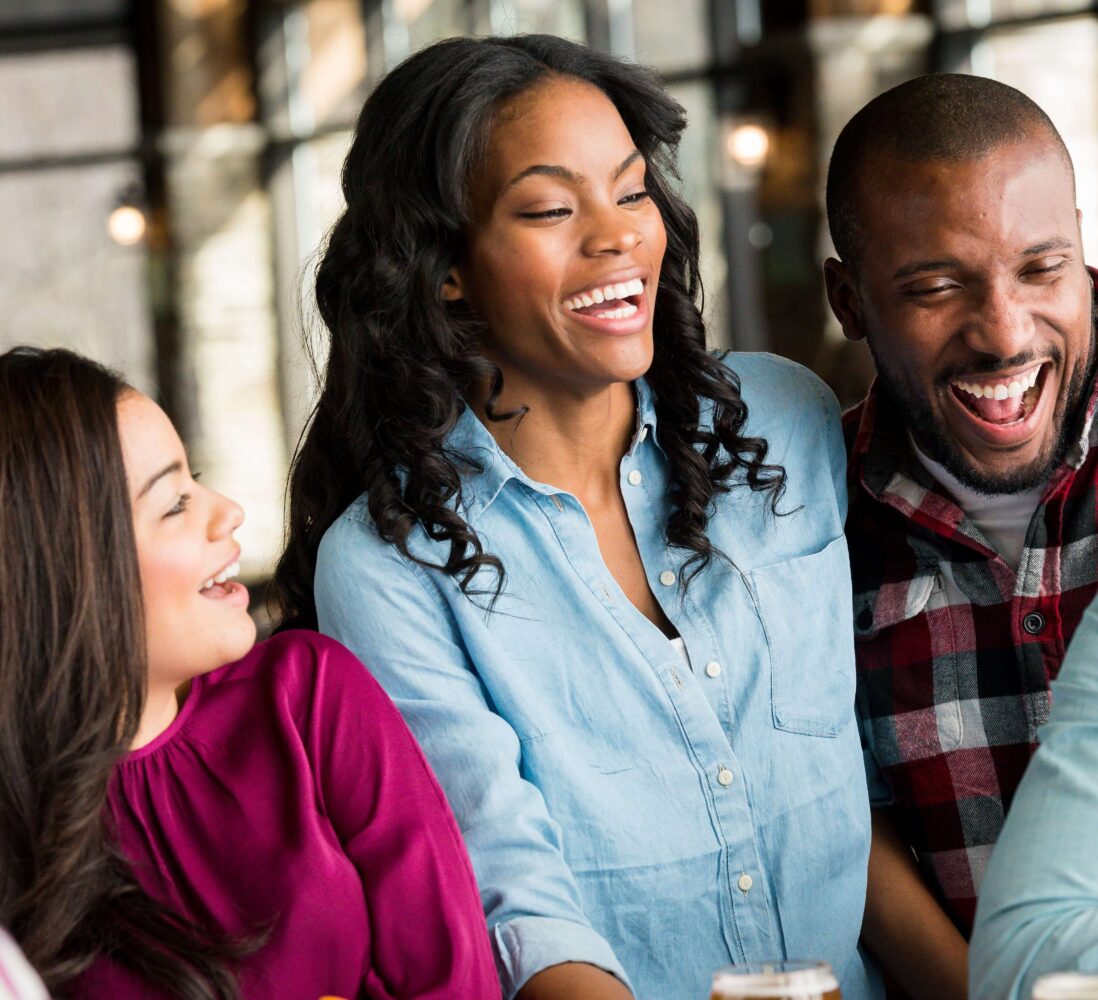 Four adults are laughing and talking together in a bright, casual indoor setting, suggesting a cheerful social gathering.