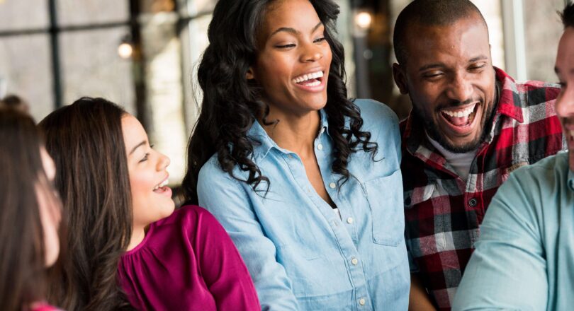 Four adults are laughing and talking together in a bright, casual indoor setting, suggesting a cheerful social gathering.