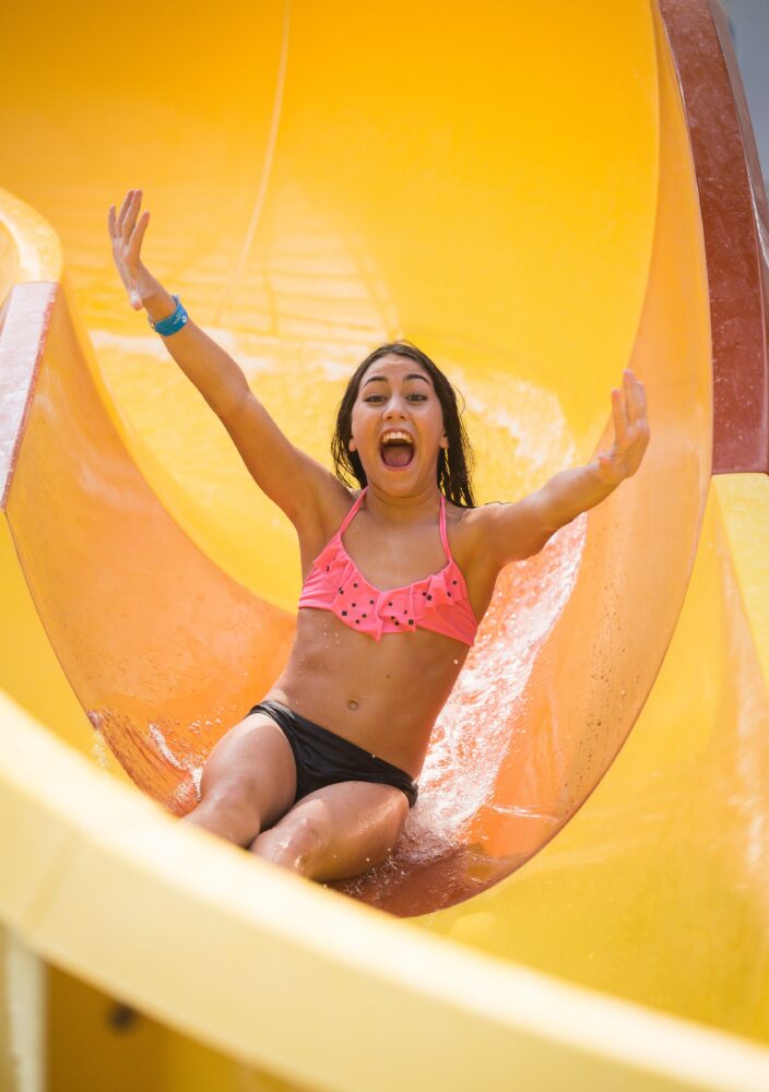 A woman in a pink bikini slides down a yellow water slide, smiling with arms raised, enjoying the ride.