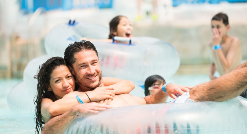 A smiling couple hugs in a pool while sitting in an inflatable tube, surrounded by children playing in the water at a water park.