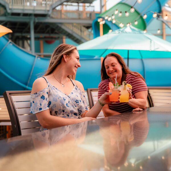 Two women smiling and clinking colorful drinks at an outdoor table, with a large blue slide and umbrellas in the background.