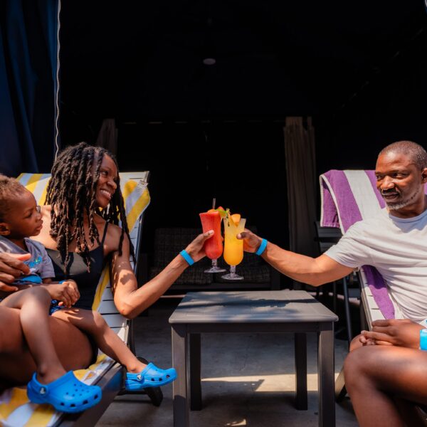 A family relaxes on lounge chairs by a pool, clinking colorful drinks and smiling together.