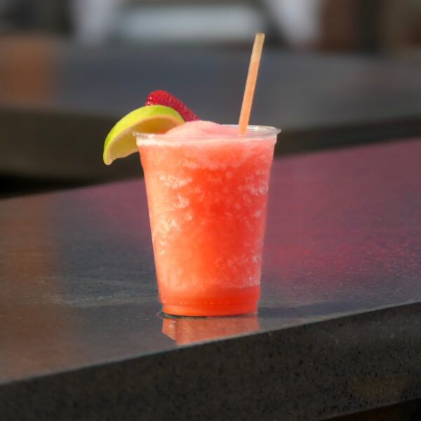 A frozen strawberry drink in a clear cup with a lime wedge and straw, sitting on a dark countertop.