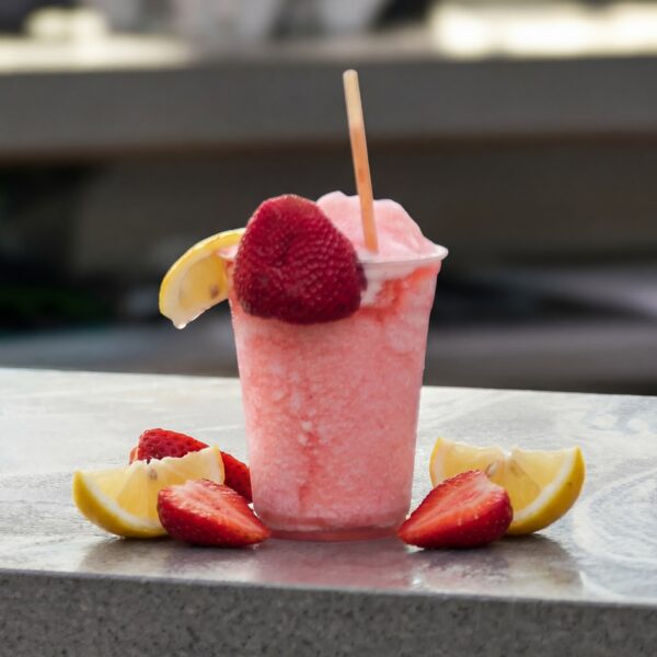A pink frozen drink in a clear cup, garnished with fresh strawberries and lemon slices, sits on a stone table.