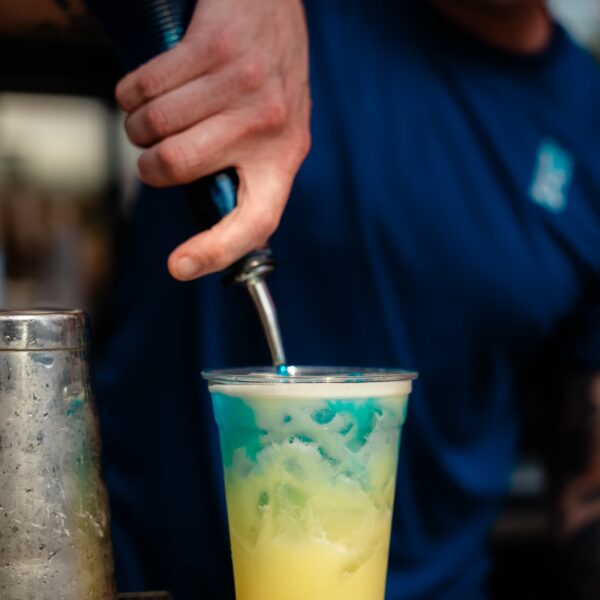 A bartender pours blue syrup into a cup of yellow cocktail, creating a layered drink. The bartender wears a blue shirt.
