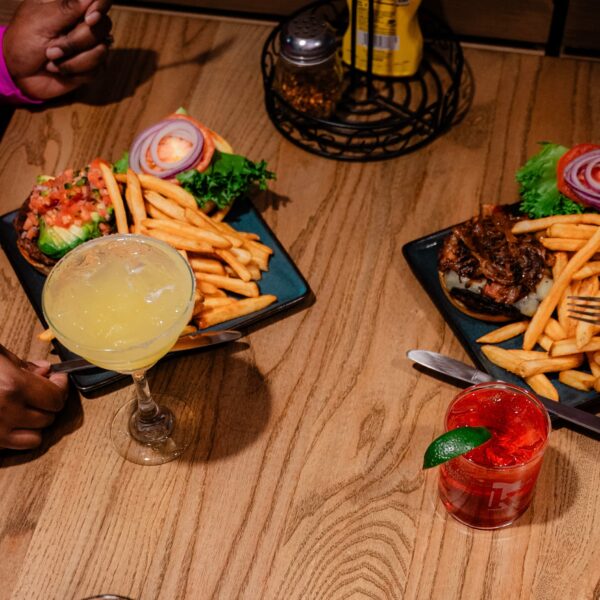 Two people eating burgers with fries, one has a yellow cocktail, the other a red drink with lime, at a wooden table in a restaurant.